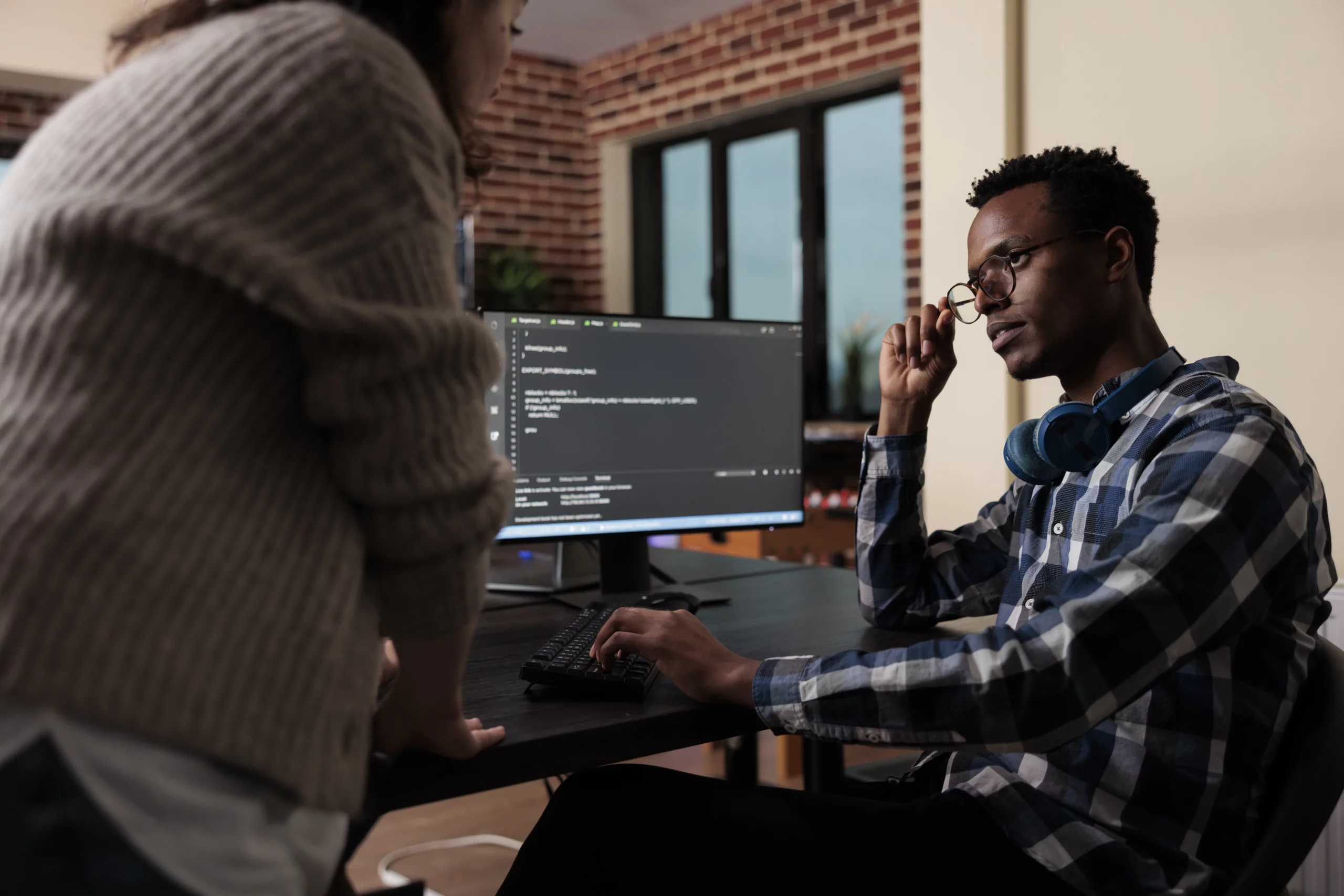 Deux développeurs web discutent d'un projet devant un écran d'ordinateur affichant du code.