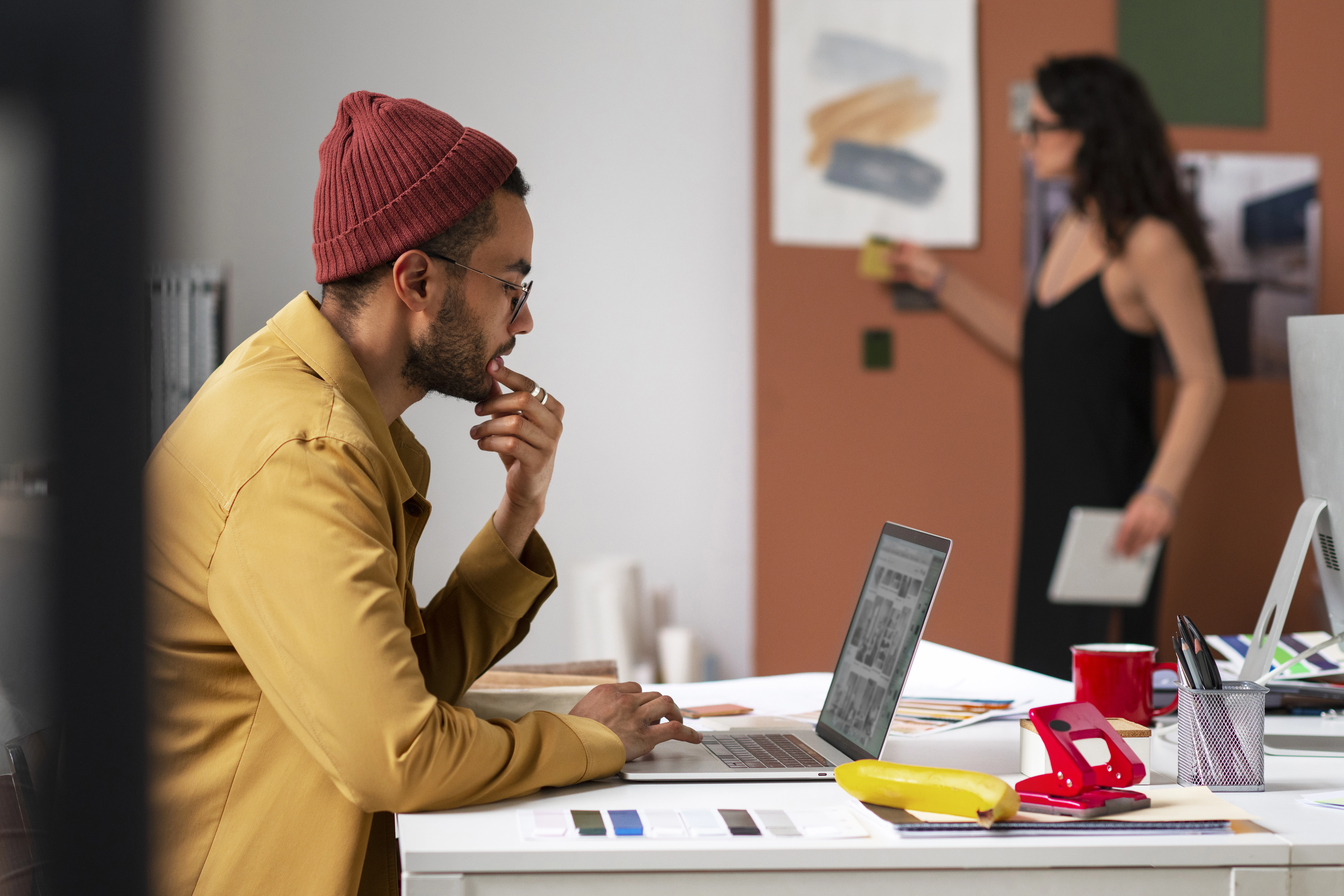 Un homme portant un bonnet rouge et une veste jaune travaille sur un ordinateur portable dans un bureau, avec une femme en arrière-plan.
