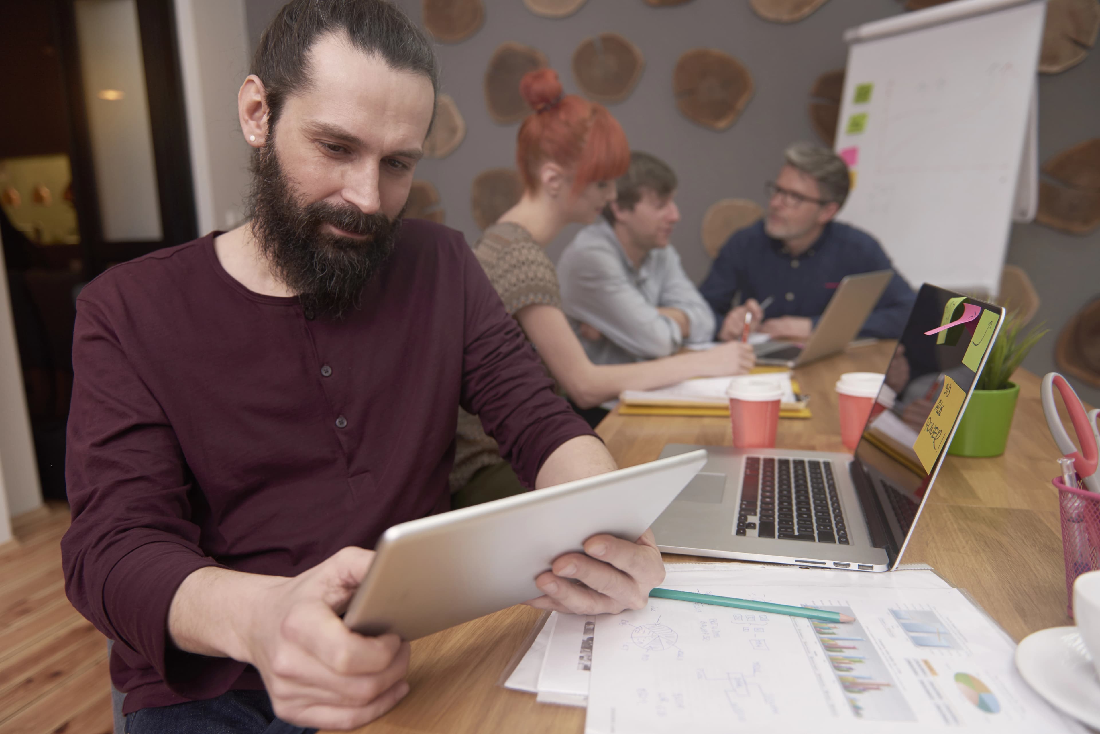 Un homme barbu utilisant une tablette lors d'une réunion de travail avec ses collègues en arrière-plan