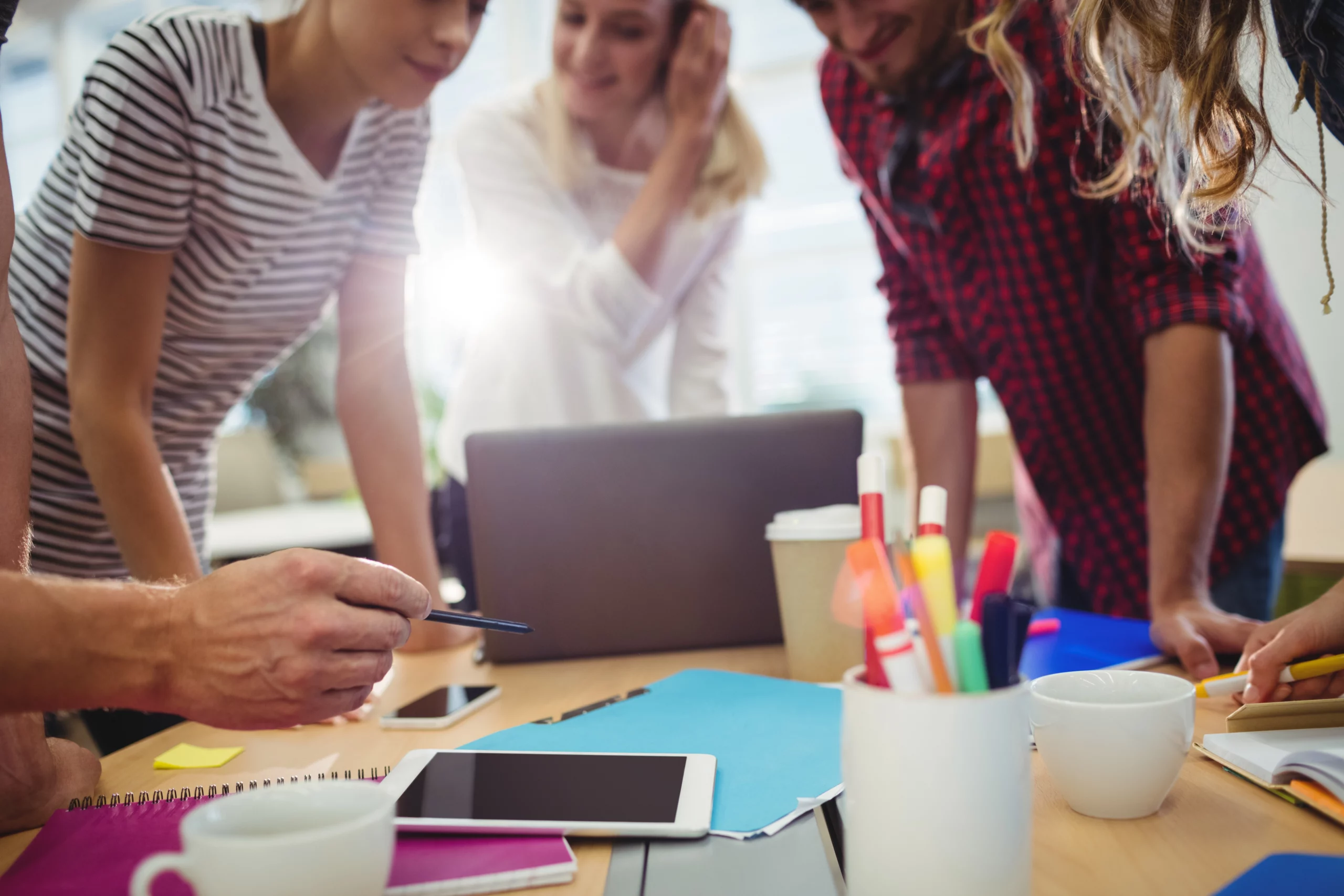Une équipe discute autour d'un ordinateur portable pour un projet digital, avec des carnets, tablettes et fournitures sur la table.